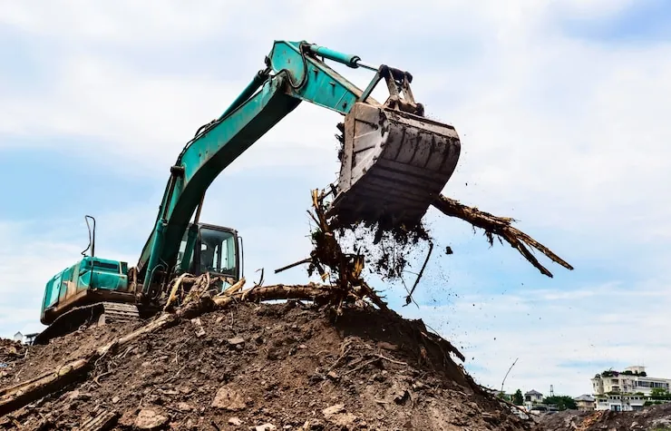 An excavator lifting a bucket of soil and roots off a heap on a plot being cleared near houses